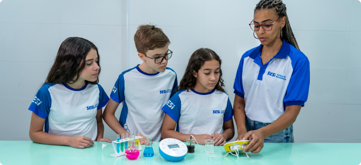 Na bancada de um laboratório de ciências, vemos uma professora, um aluno e uma aluna, todos usam óculos e luvas ao manipular tubos de ensaio com líquidos coloridos.