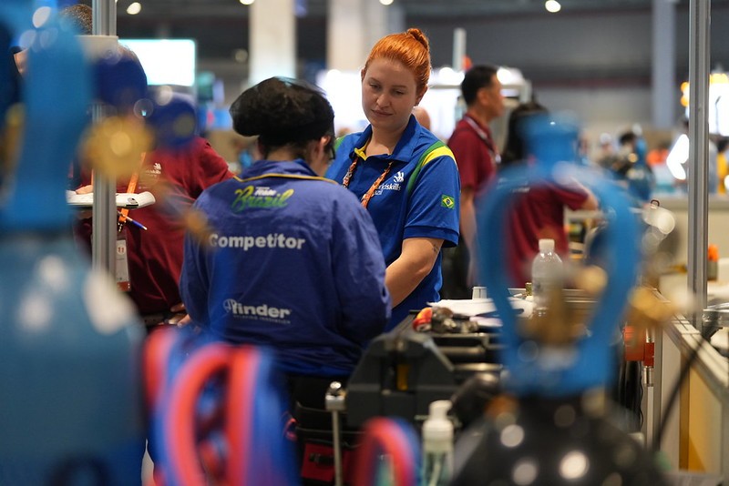 A intérprete Mariana Trapp, de cabelos ruivos, conversa com uma competidora brasileira durante a preparação para as provas da WorldSkills Shanghai 2026