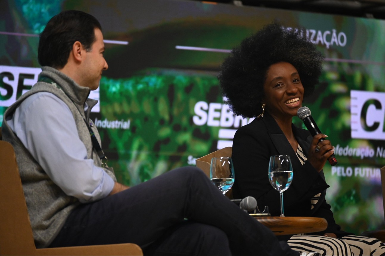 Mulher negra sorrindo com microfone ao lado de homem branco em palco com fundo verde