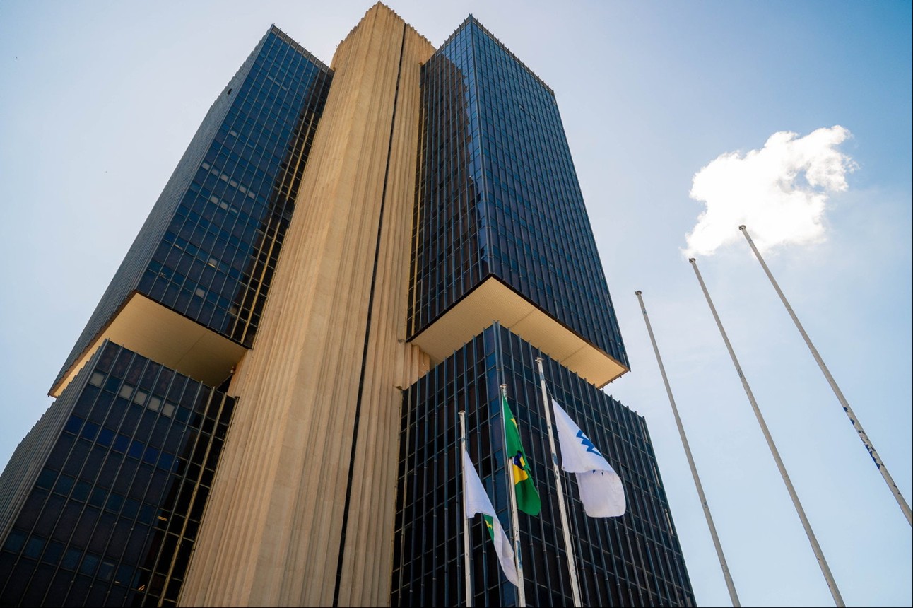 Fachada do prédio do Banco Central vista de baixo com bandeiras e céu azul ao fundo