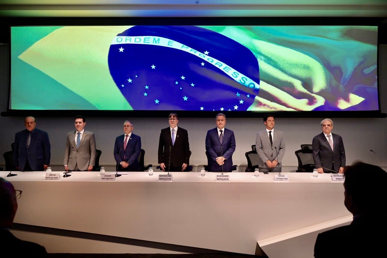 Sete homens brancos de terno em pé atrás de uma mesa com toalha branca e seus nomes com tela ao fundo com bandeira do Brasil