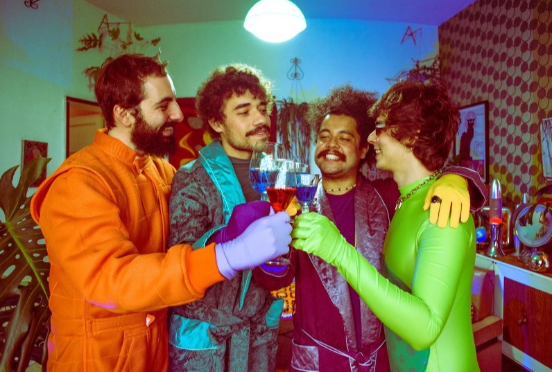 Quatro pessoas posam com taças de vinho. Cada uma, usa um macacão colorido, nas cores laranja, azul, roxo e verde