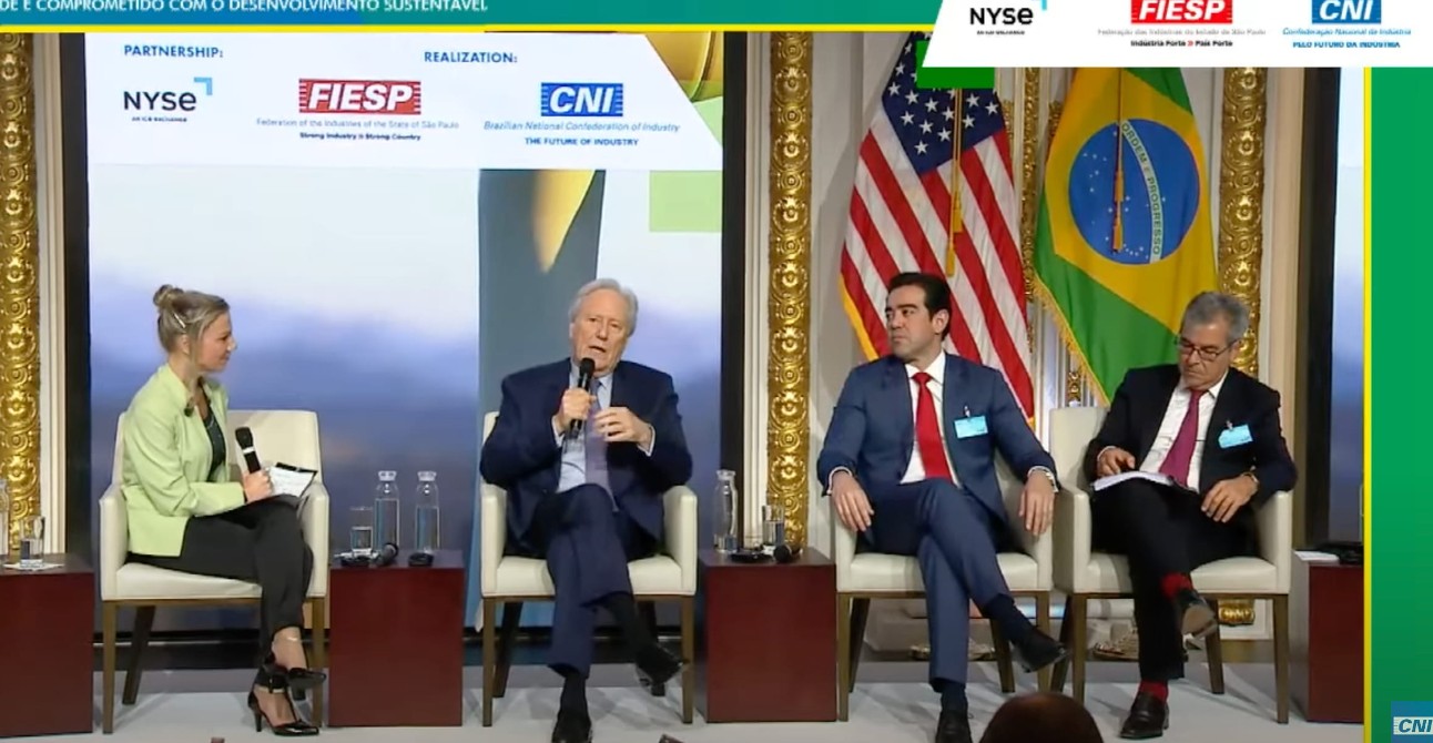 uma mulher branca e com cabelos presos, vestindo blaser verde claro, e três homens, todos de terno, estão sentados em cadeiras durante debate, com um telão e as bandeiras dos Estados Unidos e do Brasil ao fundo