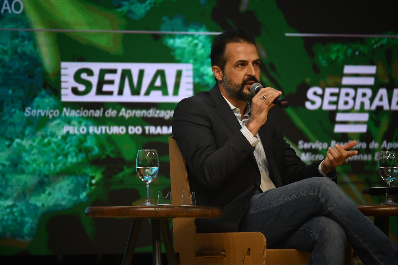 Homem branco de barca com cabelo castanho fala segurando microfone em palco sentado com tela verde com logos do SENAI e SEBRAE