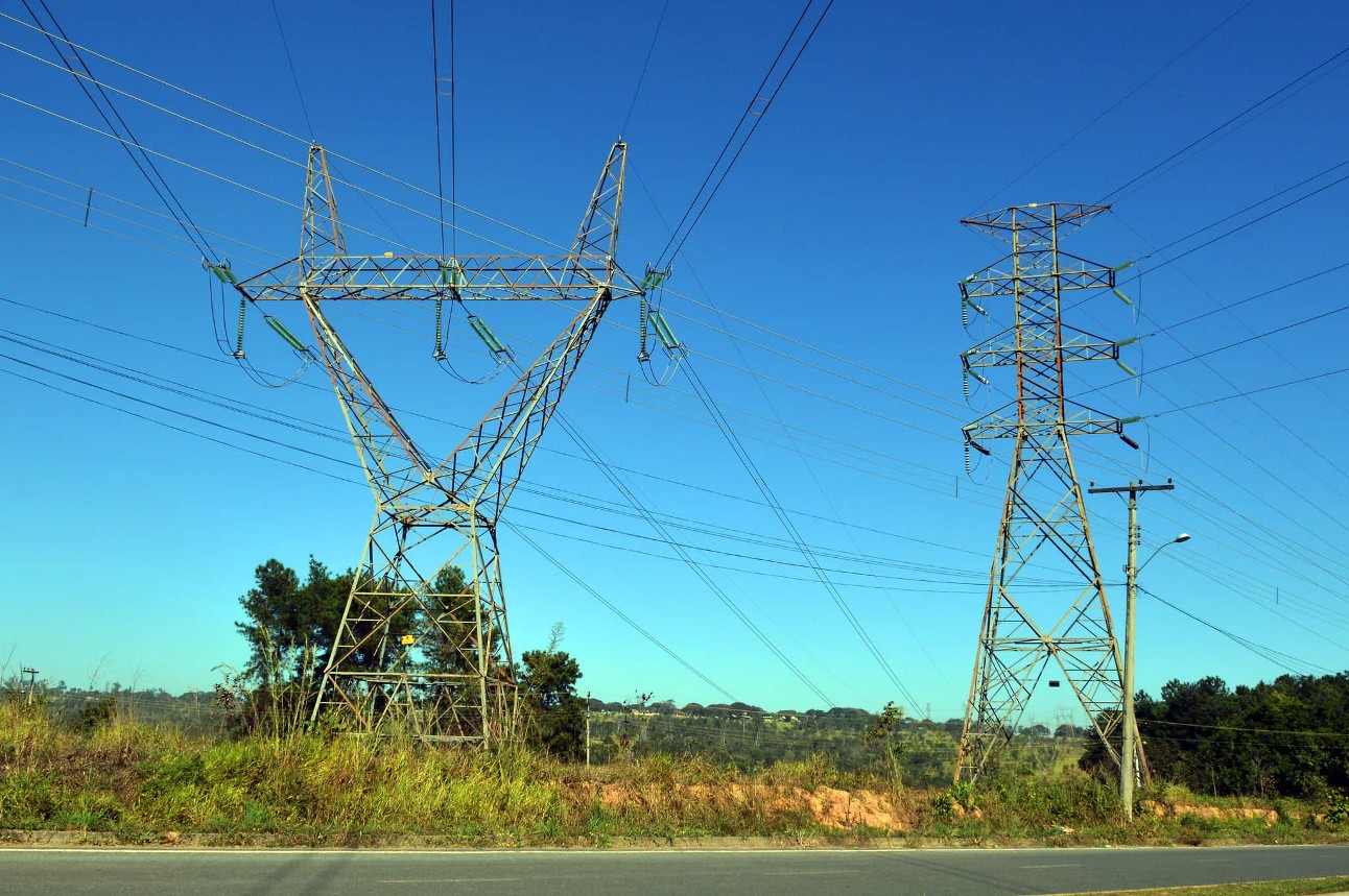 Duas torres de transmissão de energia com céu azul ao fundo, gramado e um pedaço de asfalto.