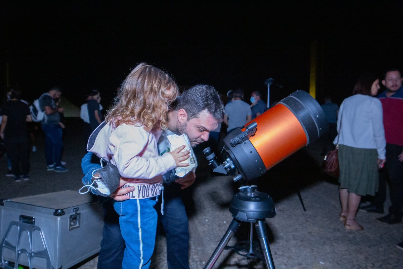 pai e filha observam telescópio. A menina é uma criança de cabelos loiros e segura uma pipoca e o homem é branco e esta abaixado para ver a constelação