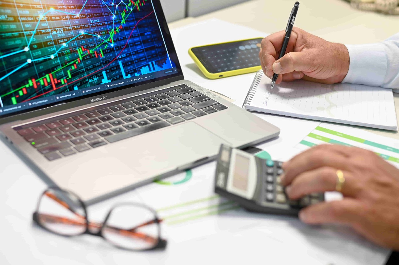 Notebook com gráfico na tela e mesa com calculadora, óculos e mãos de homem escrevendo