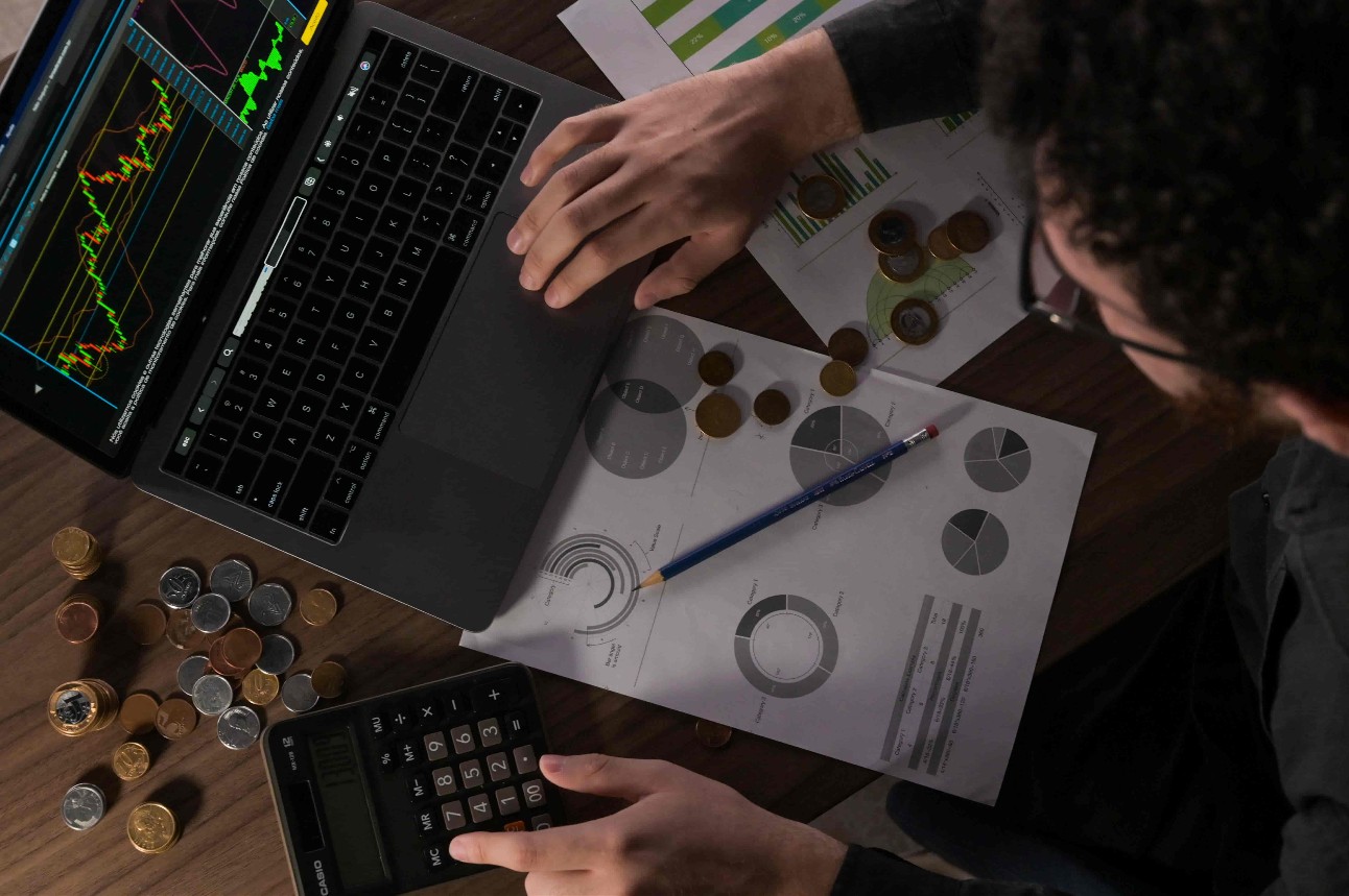 Mesa com computador com gráfico aberto, moedas e homem branco com calculadora