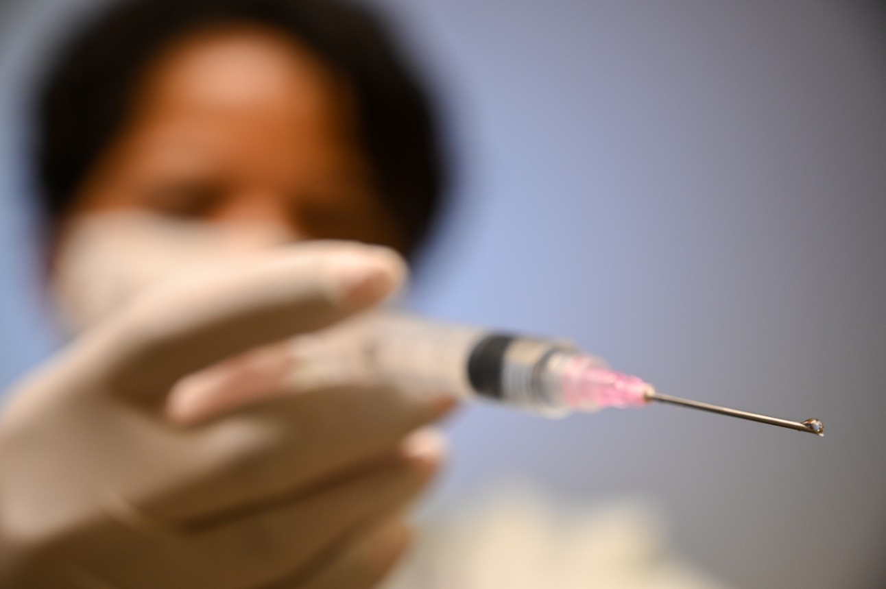 Imagem mostra enfermeira em desfoque manuseando uma seringa de vacinação. O foco da foto é a gota saindo da seringa, em primeiro plano.