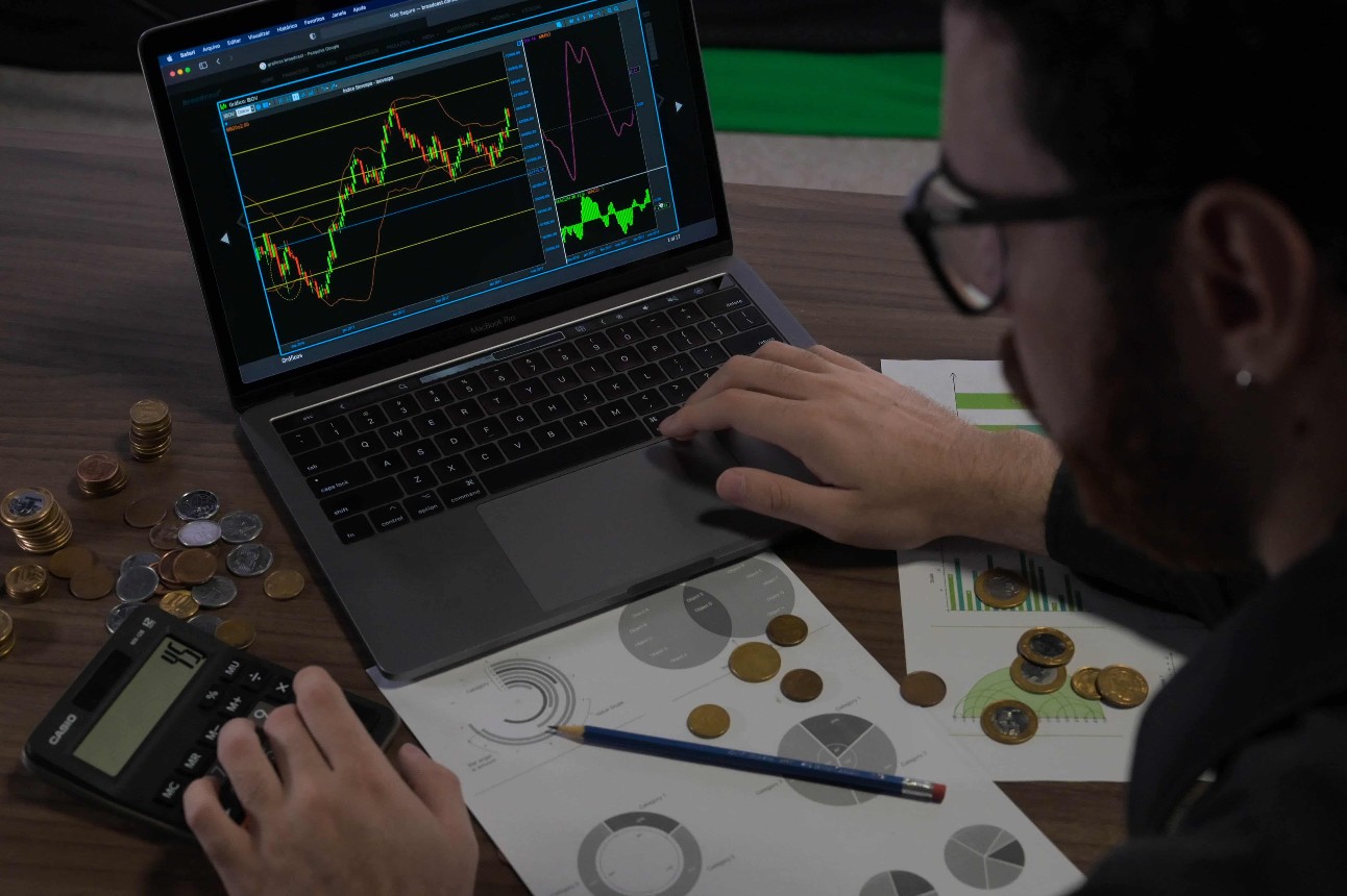 Mesa com tela de computador com gráfico, calculadora, moedas e papeis com gráficos e homem branco mexendo no computador.