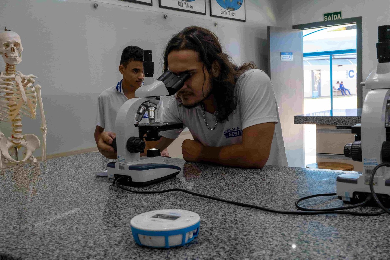 aluno branco, de cabelos longos, usa o microscópio em aula de laboratório de ciência
