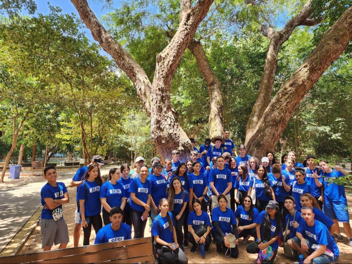 Grupo de alunos, com uniforme azul, posam em grupo de forma sorridente, em fundo com árvores