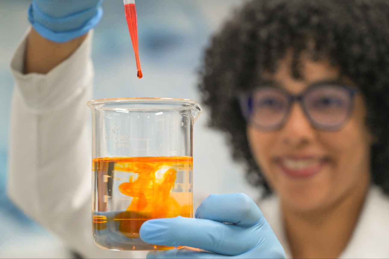 mulher negra de cabelos cacheados soltos, usando óculos e jaleco, manipula pipela e becker com líquido laranja em laboratório