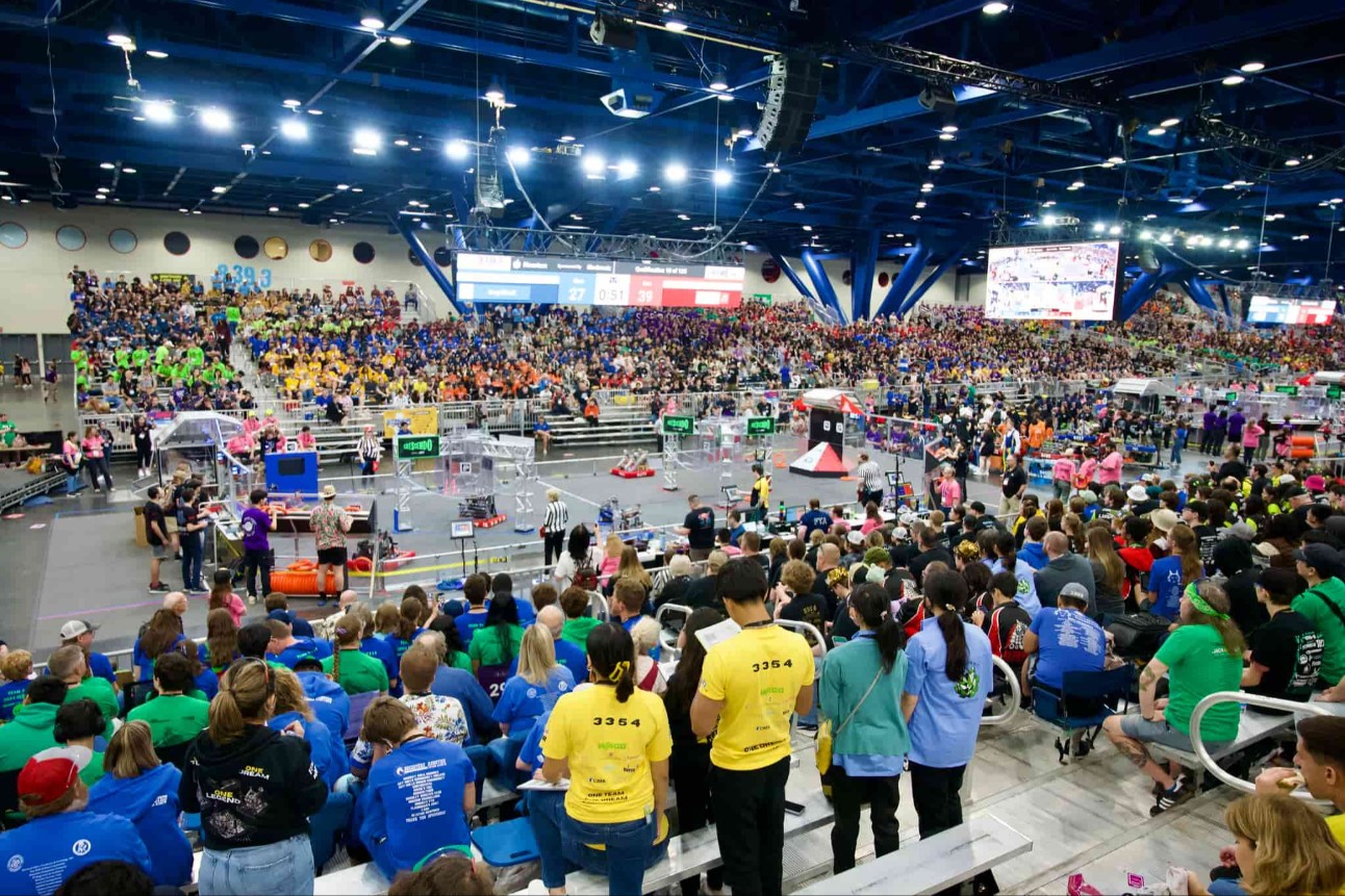 Arena lotada em Houston recebe estudantes do mundo todo para a maior competição de robótica do planeta. No centro, robôs disputam desafios enquanto torcidas vibram nas arquibancadas coloridas.