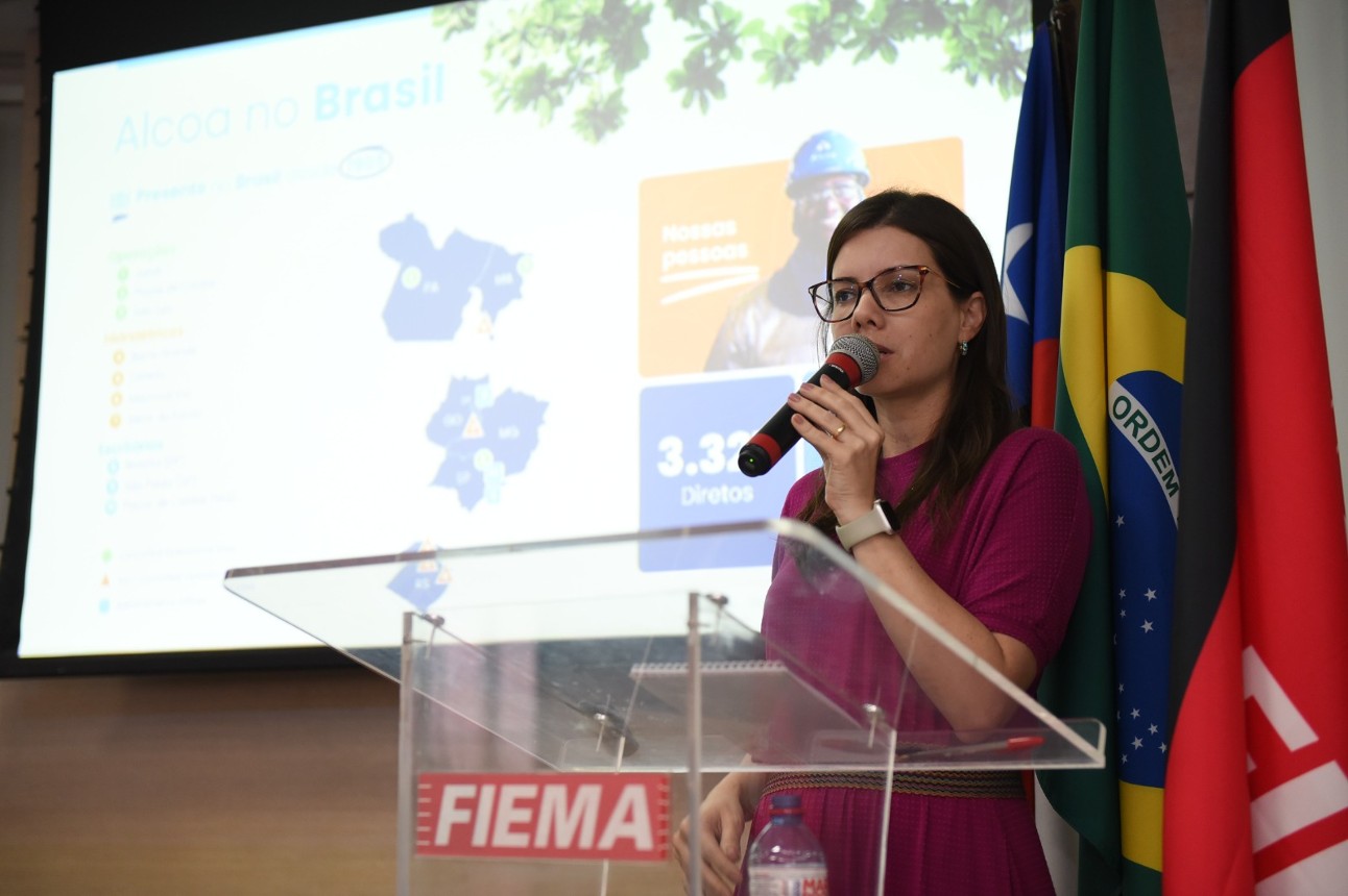 Dos quatro pilares que a Alcoa -Alumar trabalha em nível mundial, três estão associados à descarbonização da produção, tema discutido pela FIEMA mulher branca, de cabelos pretos, usa óculos e blusa vermelha fala ao microfone