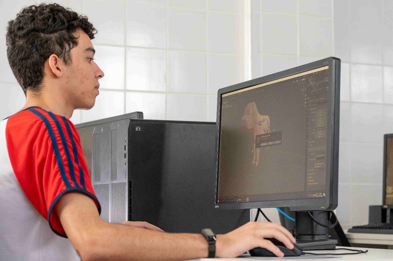 Jovem branco de camiseta branca com manga vermelha sentado em frente a computador com desenho na tela