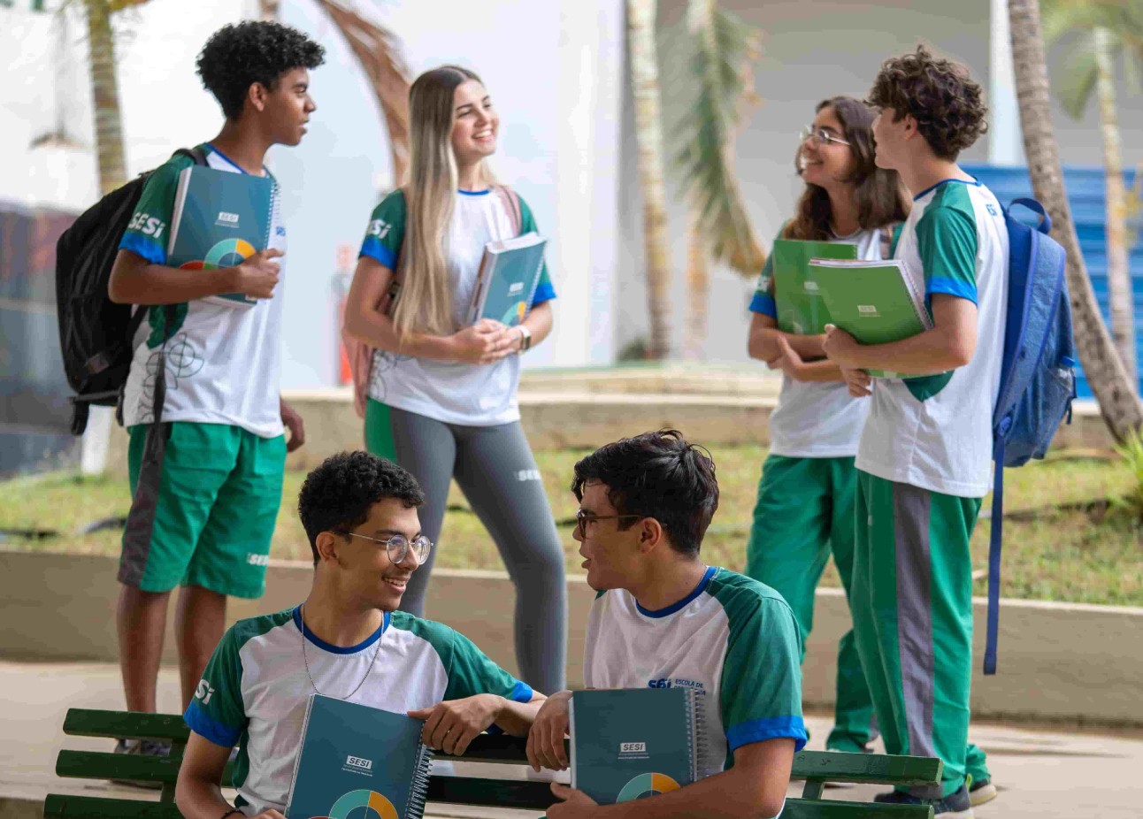 Grupo de estudantes uniformizados do SESI, sorrindo e conversando em um ambiente escolar ao ar livre, com cadernos nas mãos e rodeados por palmeiras.