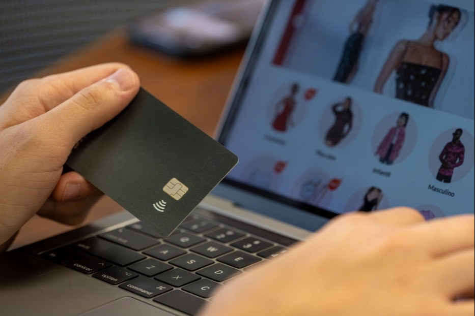 Mão de homem branco segurando cartão de crédito com tela do notebook atrás