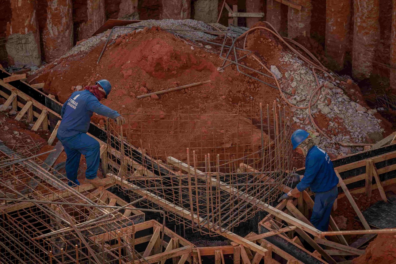 Dois homens com uniforme e capacete azul em buraco com terra e material de construção