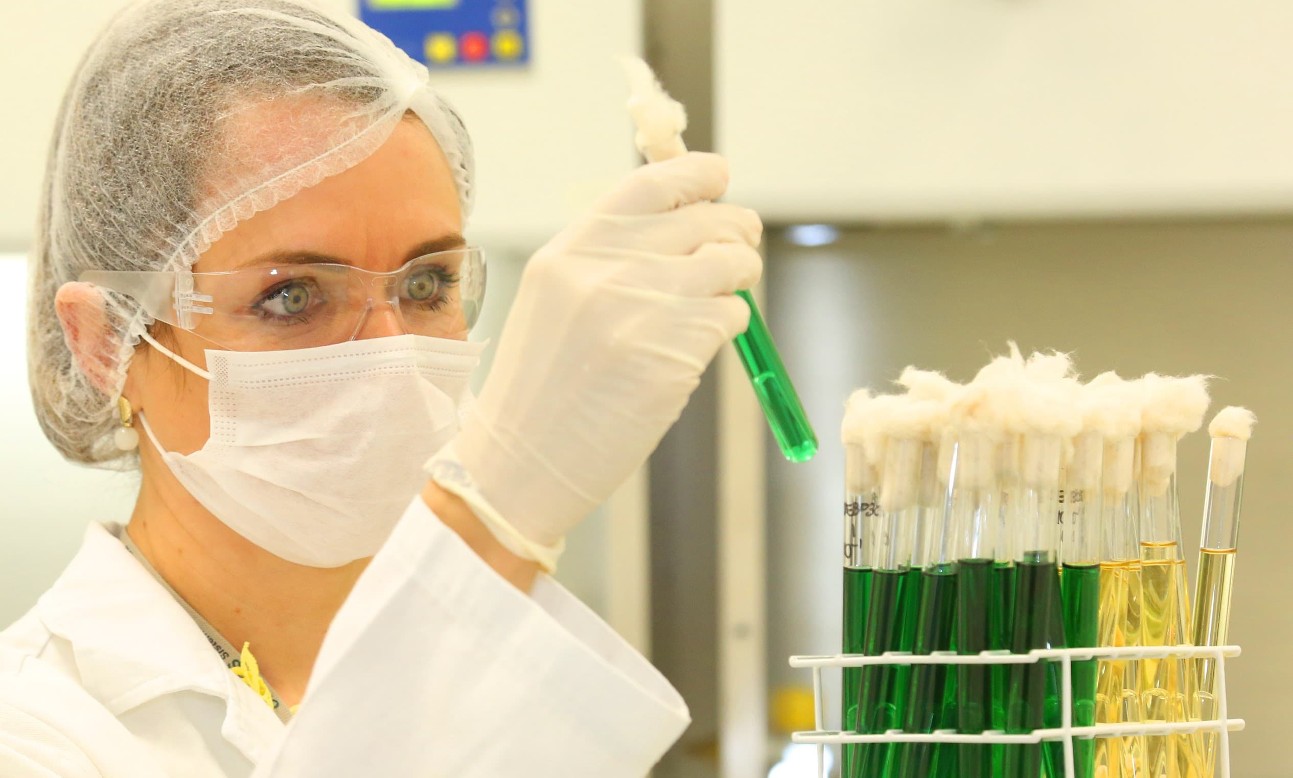 Mulher branca com touca, óculos de proteção e luva segura tubo de vidro com líquido verde em laboratório