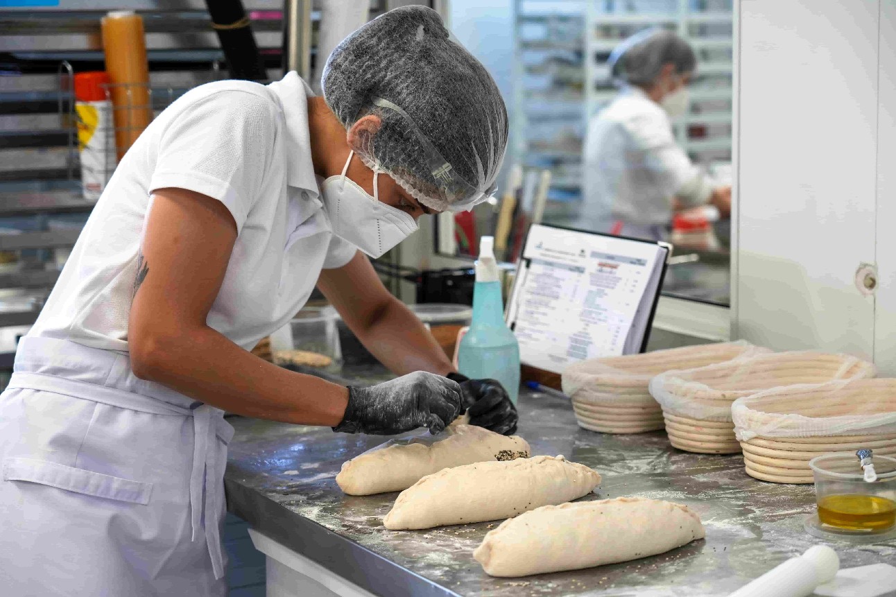 mulher, de máscara e touca de cabelo, usa avental e produz pães