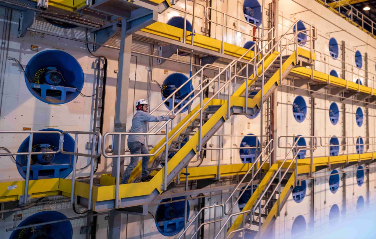 Trabalhador com capacete e protetor auricular sobe uma escada amarela em área industrial, ao lado de grandes ventiladores azuis instalados em uma parede branca. Operário sobe escada em fábrica de celulose com ventiladores industriais azuis.