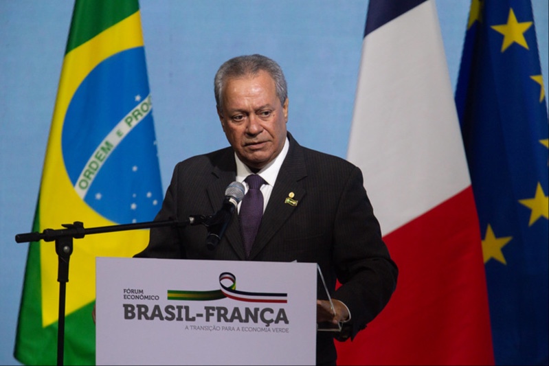 homem branco vestindo terno escuro, camisa branca e gravata vinho fala em pulplito em frente às bandeiras do Brasil e da França