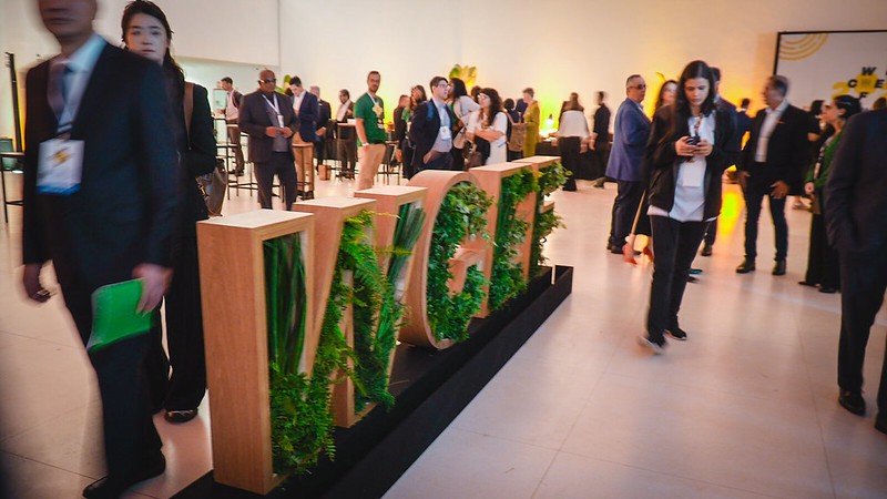 Evento corporativo elegante com pessoas em trajes sociais circulando e interagindo. No centro, um letreiro “WGF” coberto por folhagens chama atenção no ambiente moderno e iluminado.