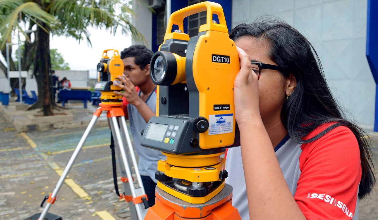 Menina com cabelo longo preto olha em equipamento amarelo. Ao fundo, menino moreno olha em equipamento semelhante