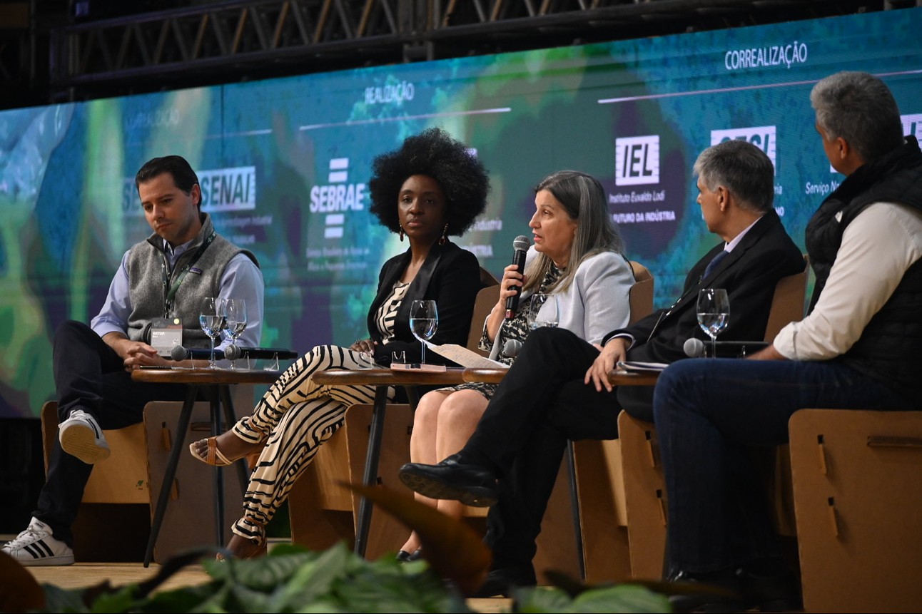 Três homens brancos, uma mulher branca e uma mulher negra sentados em palco com telão verde e azul atrás com logo do SENAI, SEBRAE e IEL