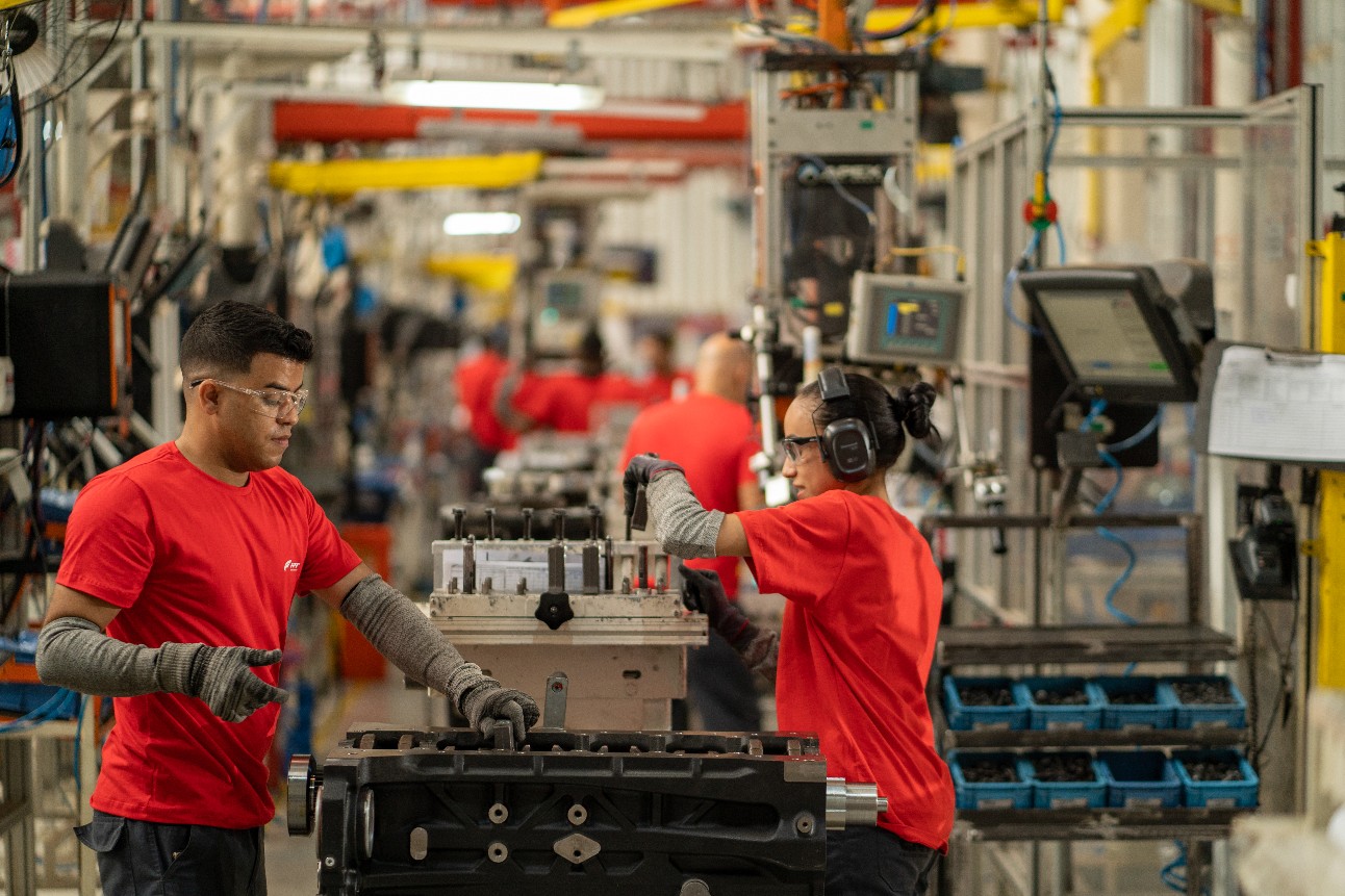 Foto colorida traz dois trabalhadores, um homem (e) e uma mulher (d), vestidos de vermelho e usando equipamentos de proteção operando equipamento industrial