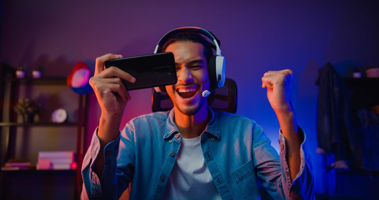 Homem feliz, jogador, usando fone de ouvido, joga videogame online com smartphone na sala de estar à noite.