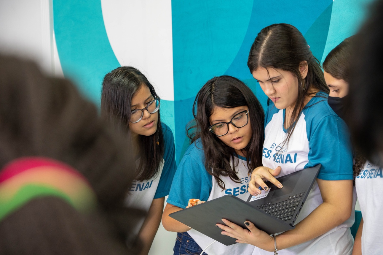quatro estudantes mulheres usam uniforme branco e azul se juntam em volta de aluna que segura um notebook