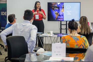 grupo de pessoas assistem palestra e, na frente, uma mulher branca, de blusa vermelha, apresenta slide