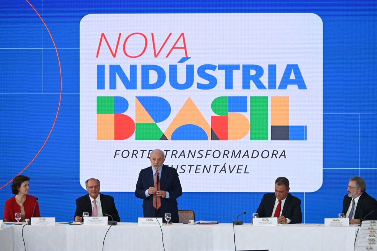 Painel digital escrito "Nova Indústria Brasil" com homem discursando de terno e pessoas sentadas ao seu lado em mesa
