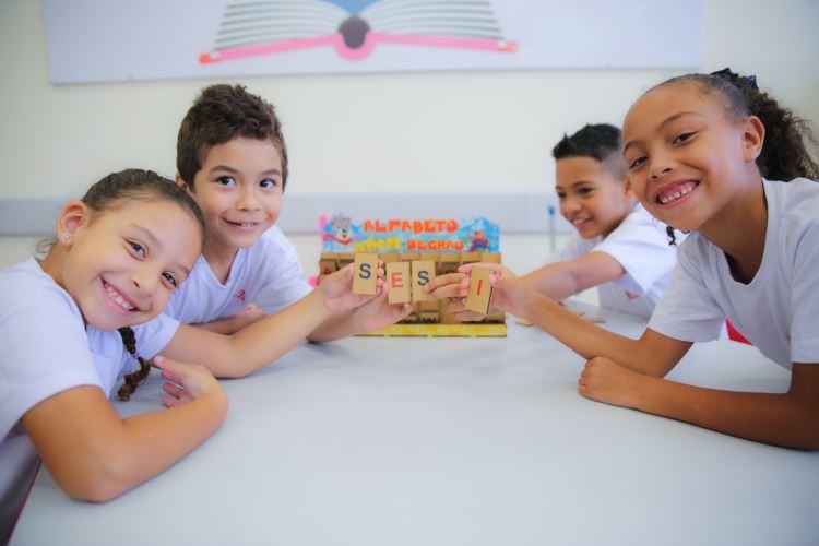 Quatro crianças, duas negras e duas brancas, dois meninos e duas meninas, posam para foto com sorriso no rosto na sala de aula. As crianças estão sentadas numa mesa e seguram um brinquedo educacional