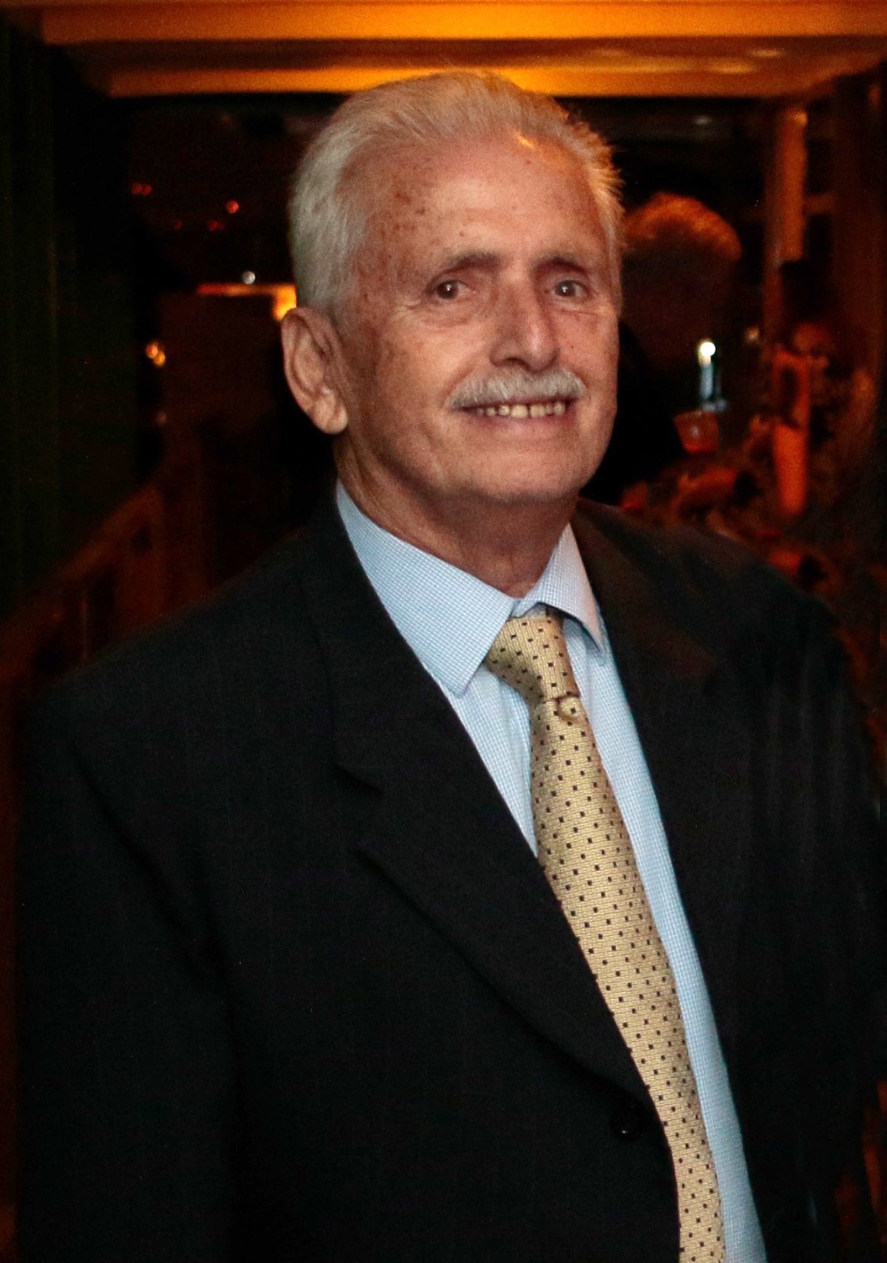 homem branco, idoso, de cabelo e bigodes brancos, usa terno escuro, camisa branca e gravata dourada em um evento