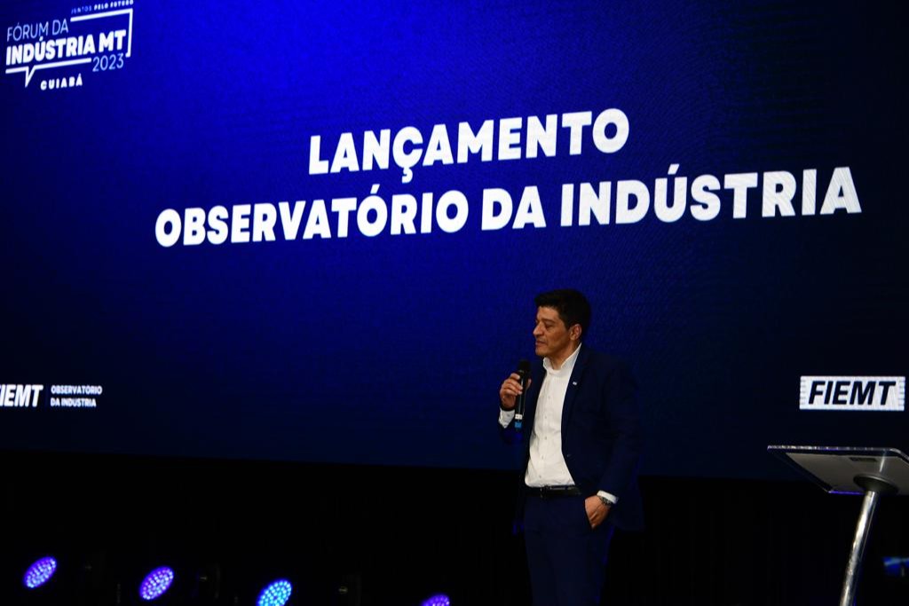 Marcelo Bispo, gerente do Observatório Nacional da Indústria, apresenta o lançamento do Observatório em evento presencial. Ele usa terno preto e branco e está atrás de um telão azul.