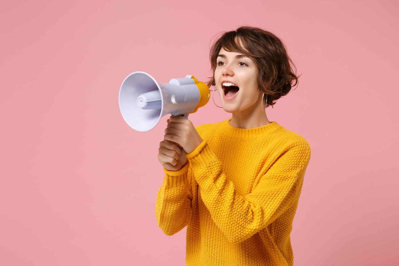 Menina alegre jovem, de cabelos castanhos, com suéter amarelo posando em estúdio de fundo rosa pastel, gritando ao microfone