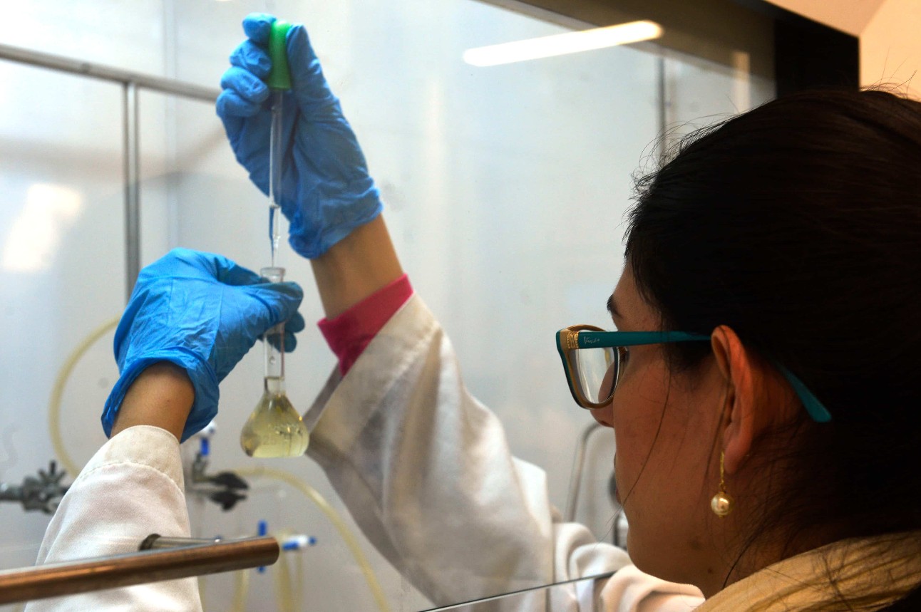 Mulher de costas em laboratório com luvas azuis segurando vidro com líquido dentro