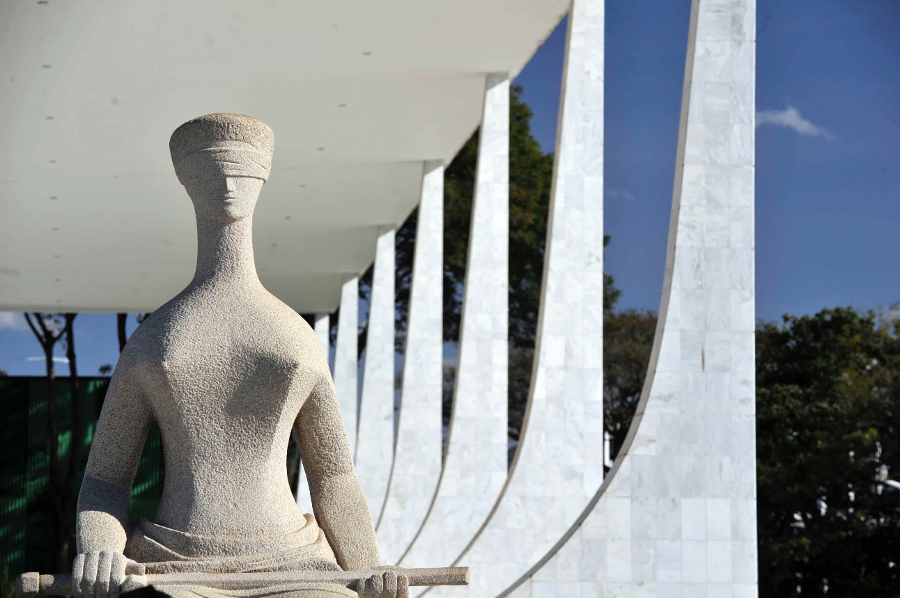 Foto colorida de estátua de pedra clara de mulher vendada representando a justiça, em frente às colunas brancas do prédio do STF em Brasíia