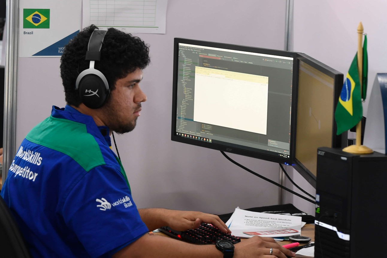 homem branco, com camiseta azul, usa fone de ouvido preto e usa o computador