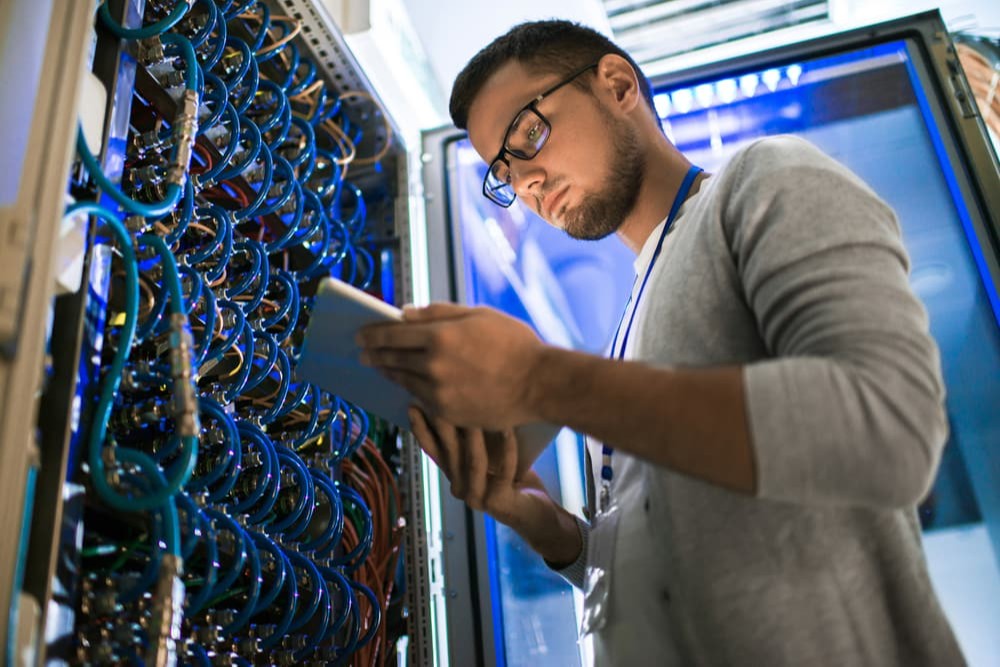 Imagem: Retrato de homem jovem moderno segurando laptop em pé na sala do servidor trabalhando com supercomputador em luz azul