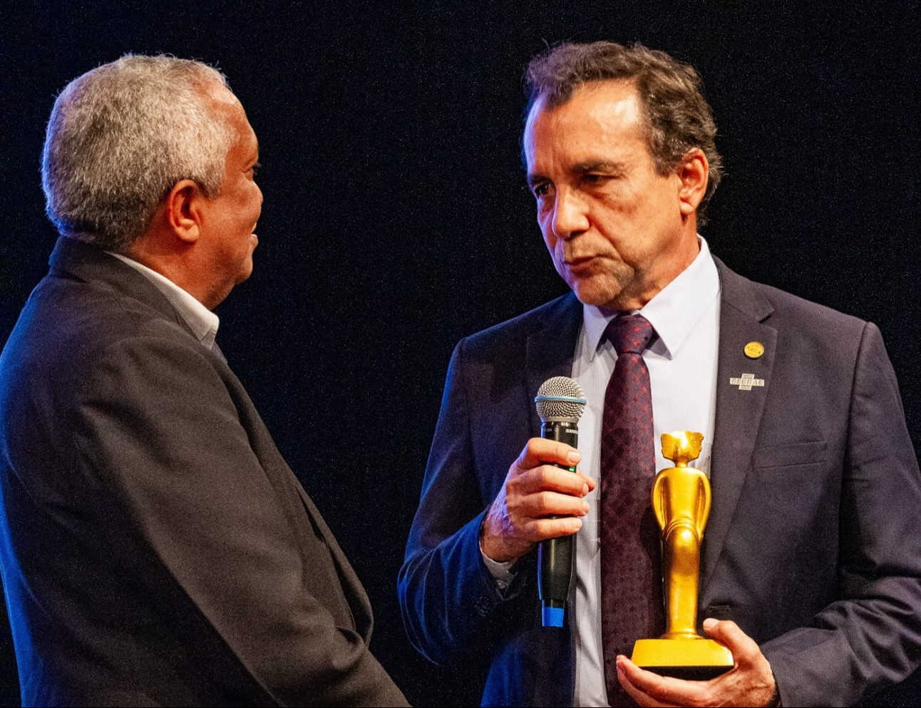 imagem colorida de homem entregando prêmio a outro homem, enquanto segura um microfone