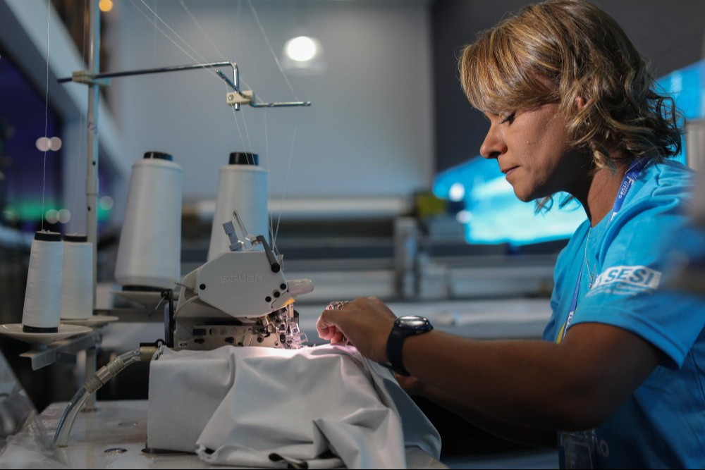Mulher loira em máquina de costura com tecido branco e usando camiseta azul com logo do SESI