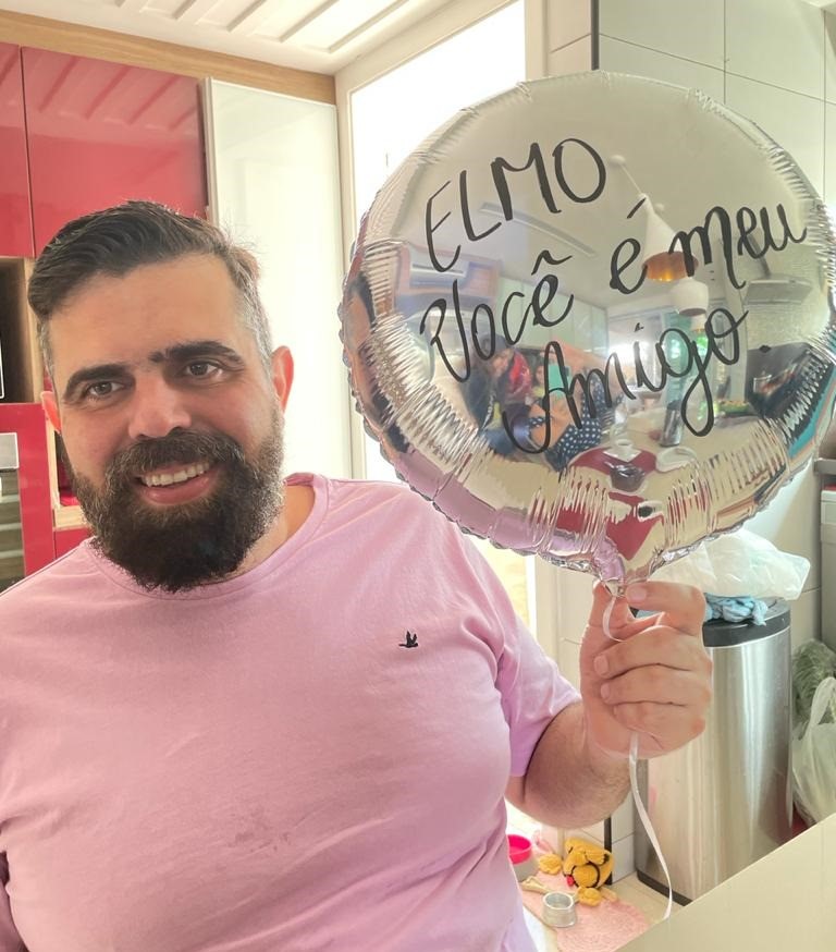 Foto:  Homem, de blusa branca, segurando um balão cinza, que tem escrito "Elmo, você é meu amigo".