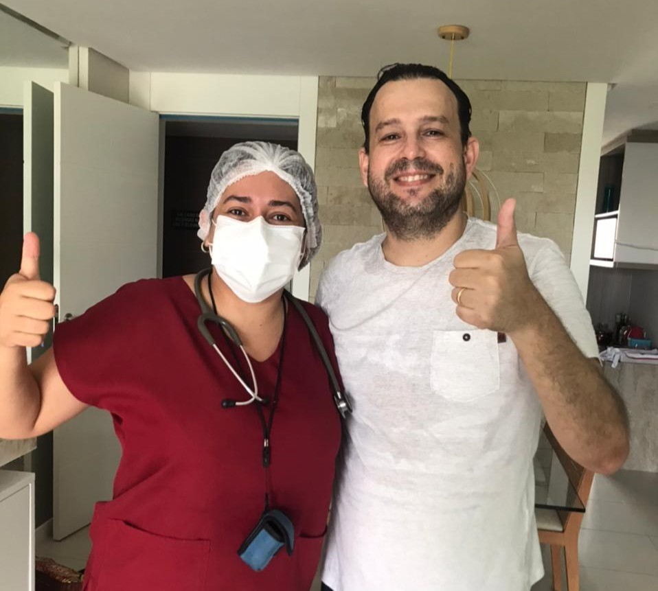 Foto: enfermeira de máscara branca e macacão vermelho, está abraçando homem alto , sorridente, de blusa branca. Ambos fazem o sinal de "joinha" com a mão.