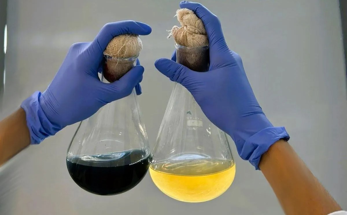 Dois vidros com líquido escuro e outro amarelo são segurados por mãos com luvas azuis