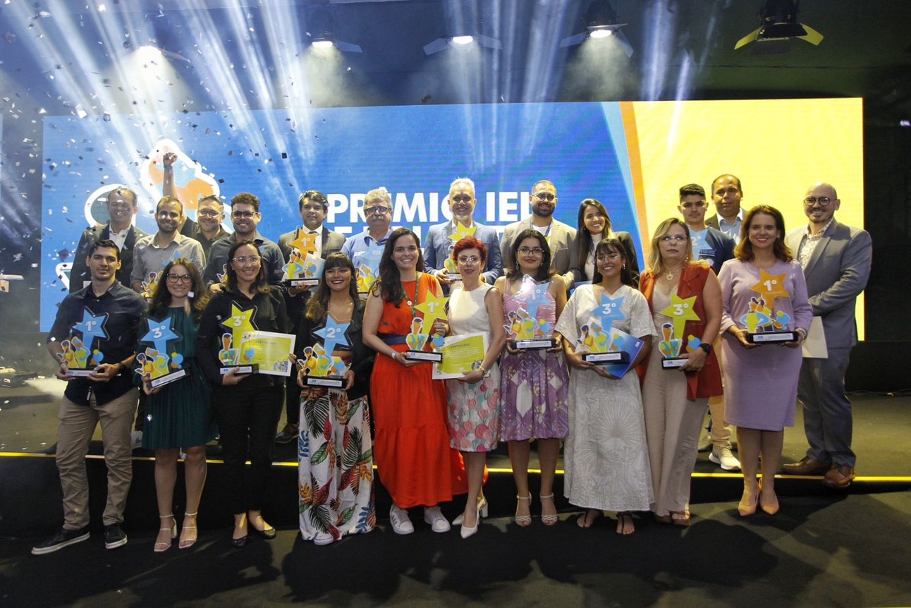 Pessoas diversas em pé segurando prêmios nas mãos com fundo "Prêmio IEL"