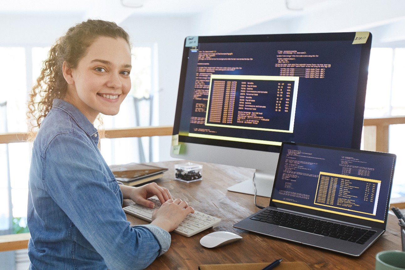 Retrato de uma desenvolvedora de TI sorrindo para a câmera enquanto digita no teclado com o código de programação preto e laranja na tela do computador e laptop no interior do escritório